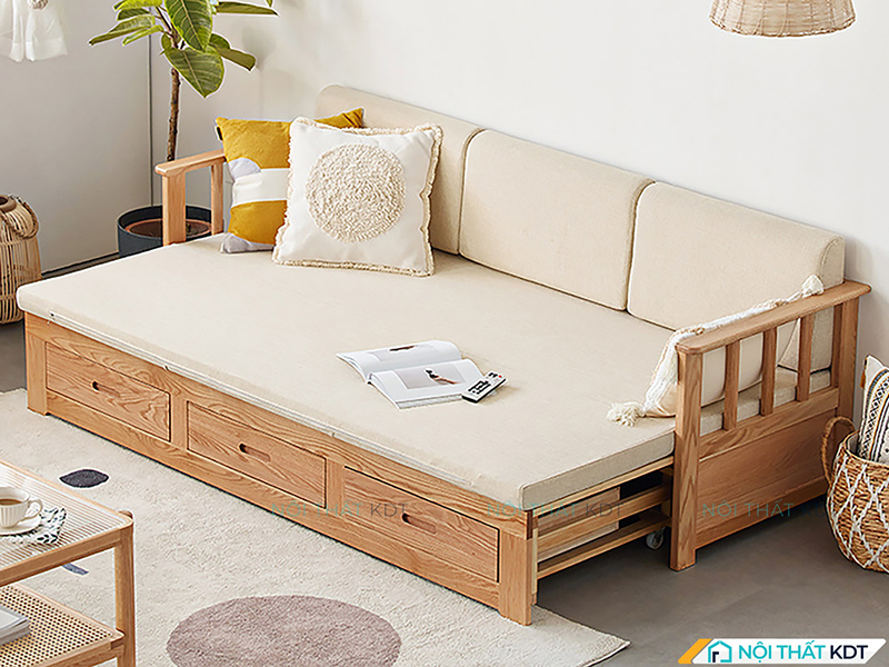 Sofa giường đa năng khung gỗ tự nhiên kê sát tường với ngăn kéo lưu trữ bên dưới, đệm màu kem kết hợp gối trang trí vàng và trắng tạo điểm nhấn